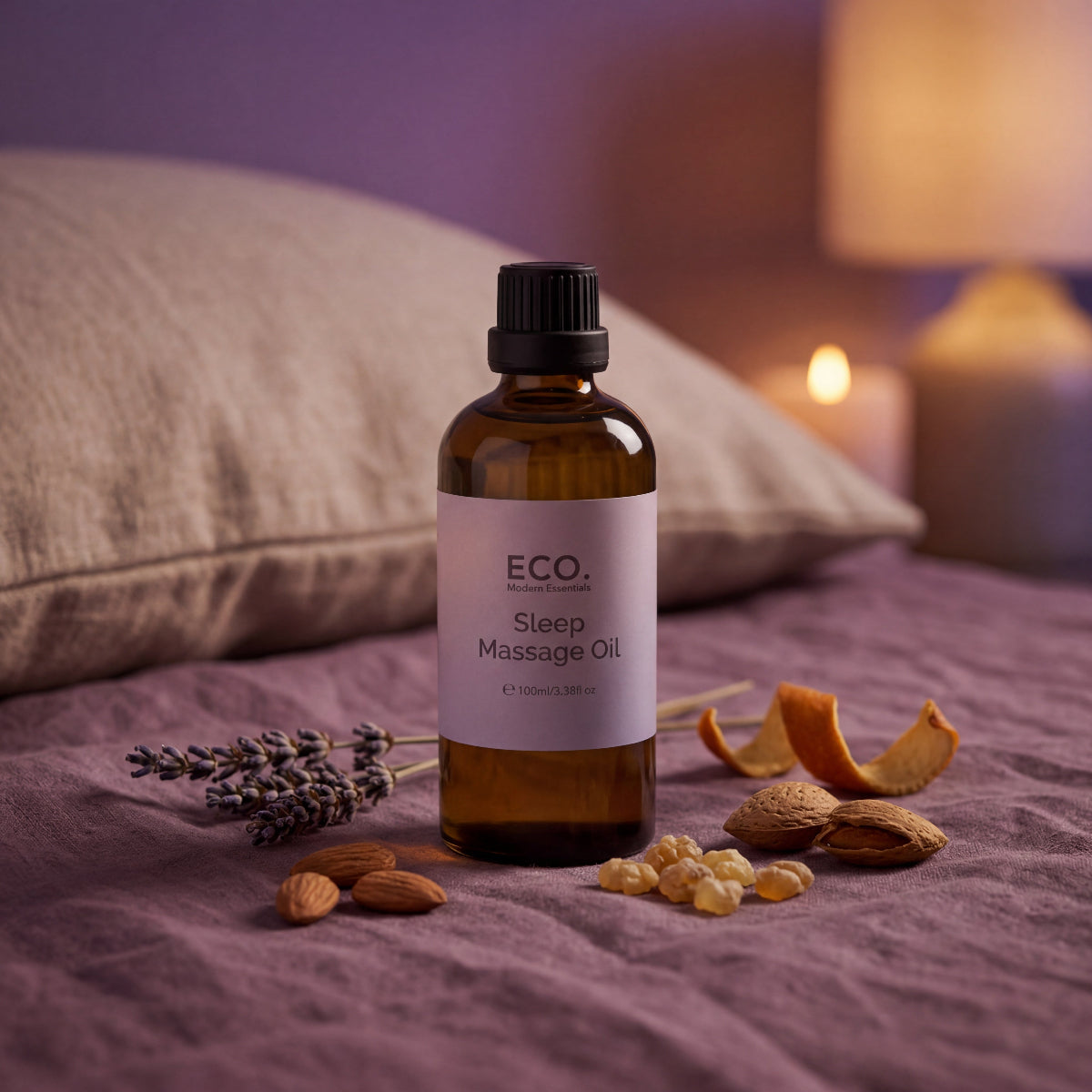 Bottle of ECO Sleep Massage Oil on a bed with lavender, almonds, and a lit candle in the background.
